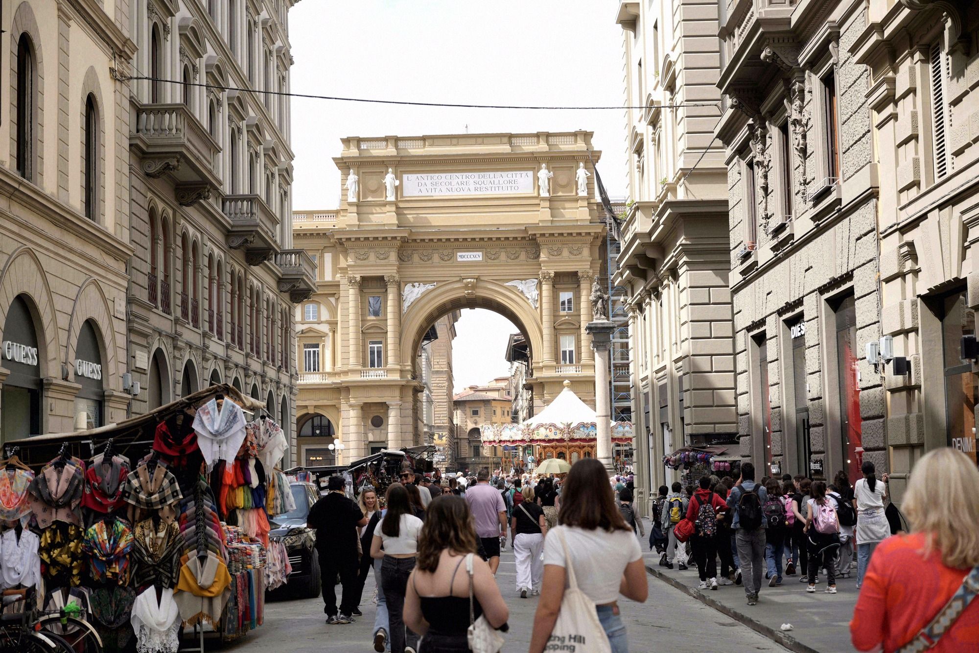Belebte Einkaufsstrasse in Richtung Piazza della Repubblica mit dem charakteristischen Triumphbogen im Hintergrund und Strassenhaendlern im Vordergrund.