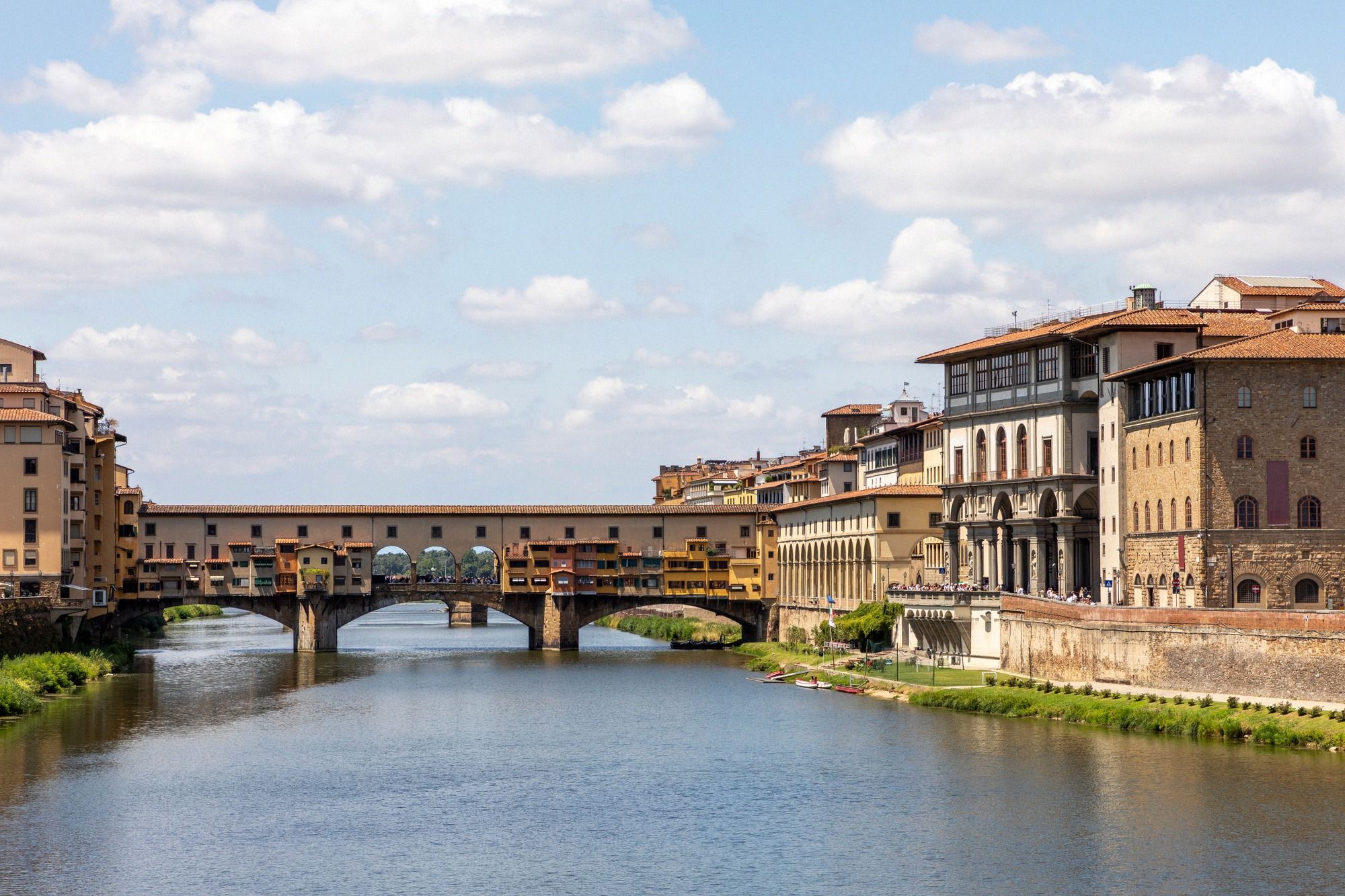 Die historische Ponte Vecchio bei sonnigem Sommertaglicht mit den charakteristischen Botteghe (Läden) und dem Arno darunter.