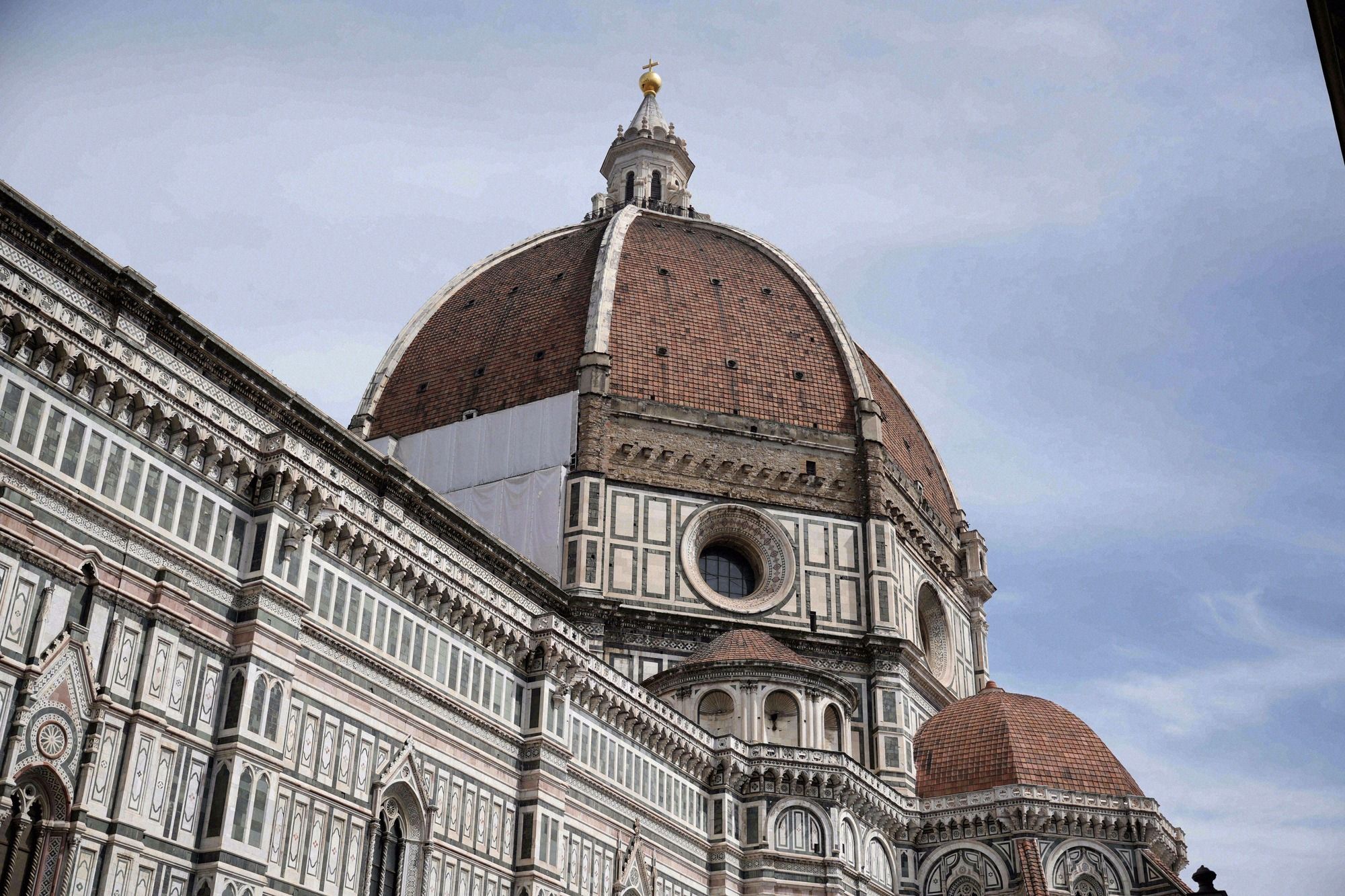Eindrucksvolle Nahaufnahme der Brunelleschi-Kuppel des Florentiner Doms mit dem goldenen Kreuz auf der Laterne und dem gruenweissen Marmordekor des Obergadens.