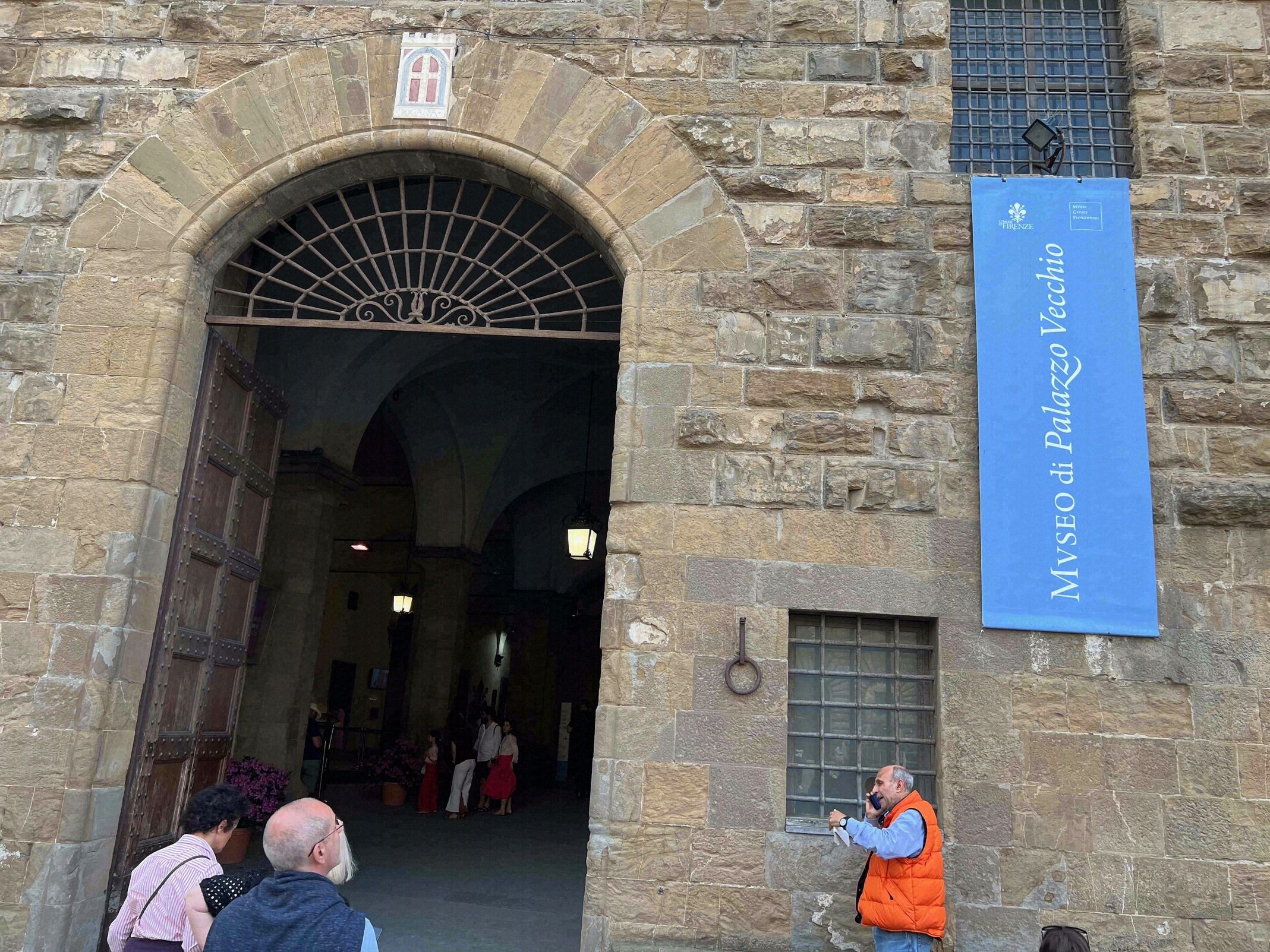 Rustikaler Eingangsportal des Palazzo Vecchio aus Sandstein mit dem blauen Schild zum Museo di Palazzo Vecchio und dem Florentiner Lilienwappen ueber dem Bogen.