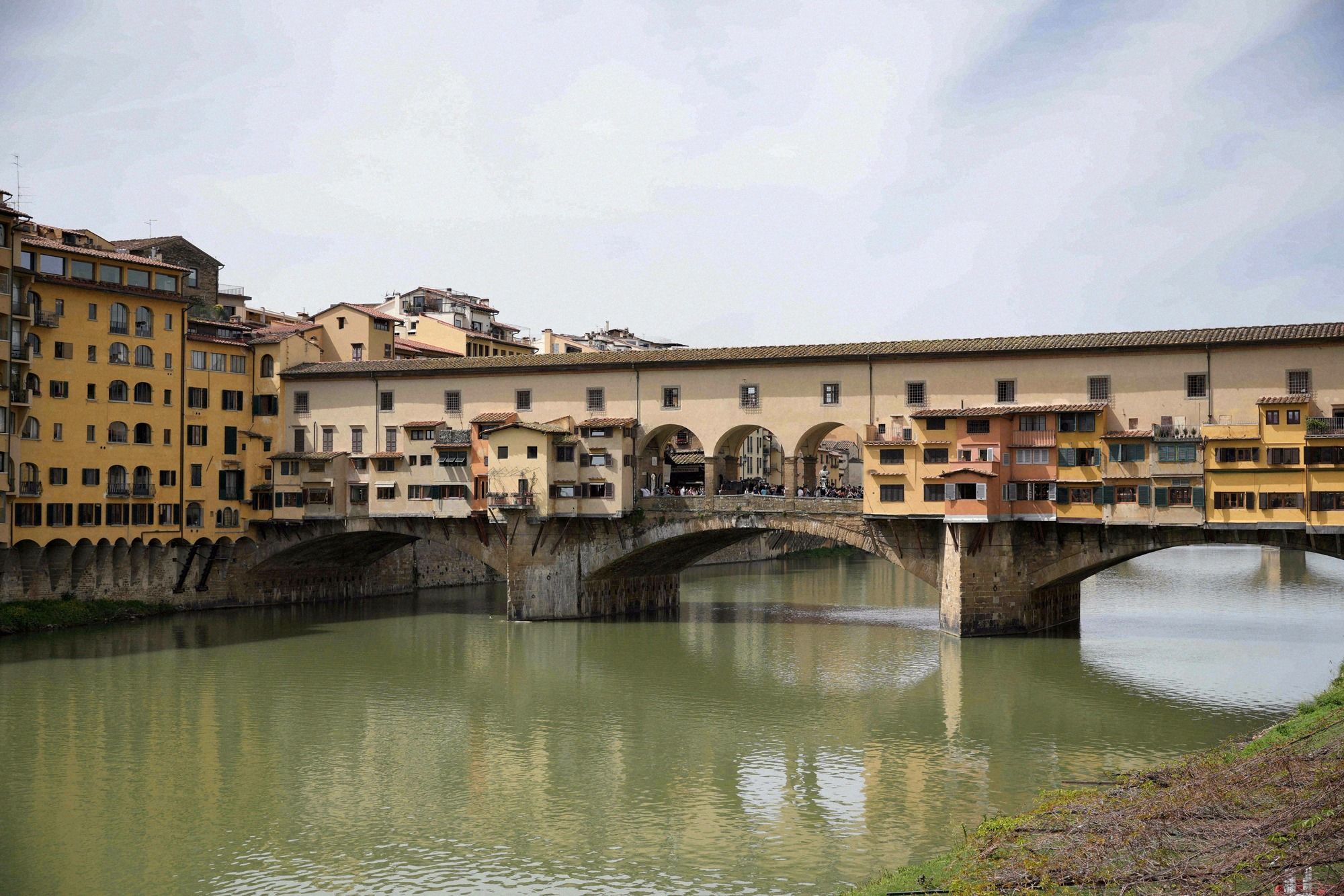 Der Ponte Vecchio ueber den Arno aus der Naeher, mit den charakteristischen ockerfarbenen Haeuseraufbauten und dem Spiegelbild der Bruecke im Fluss.