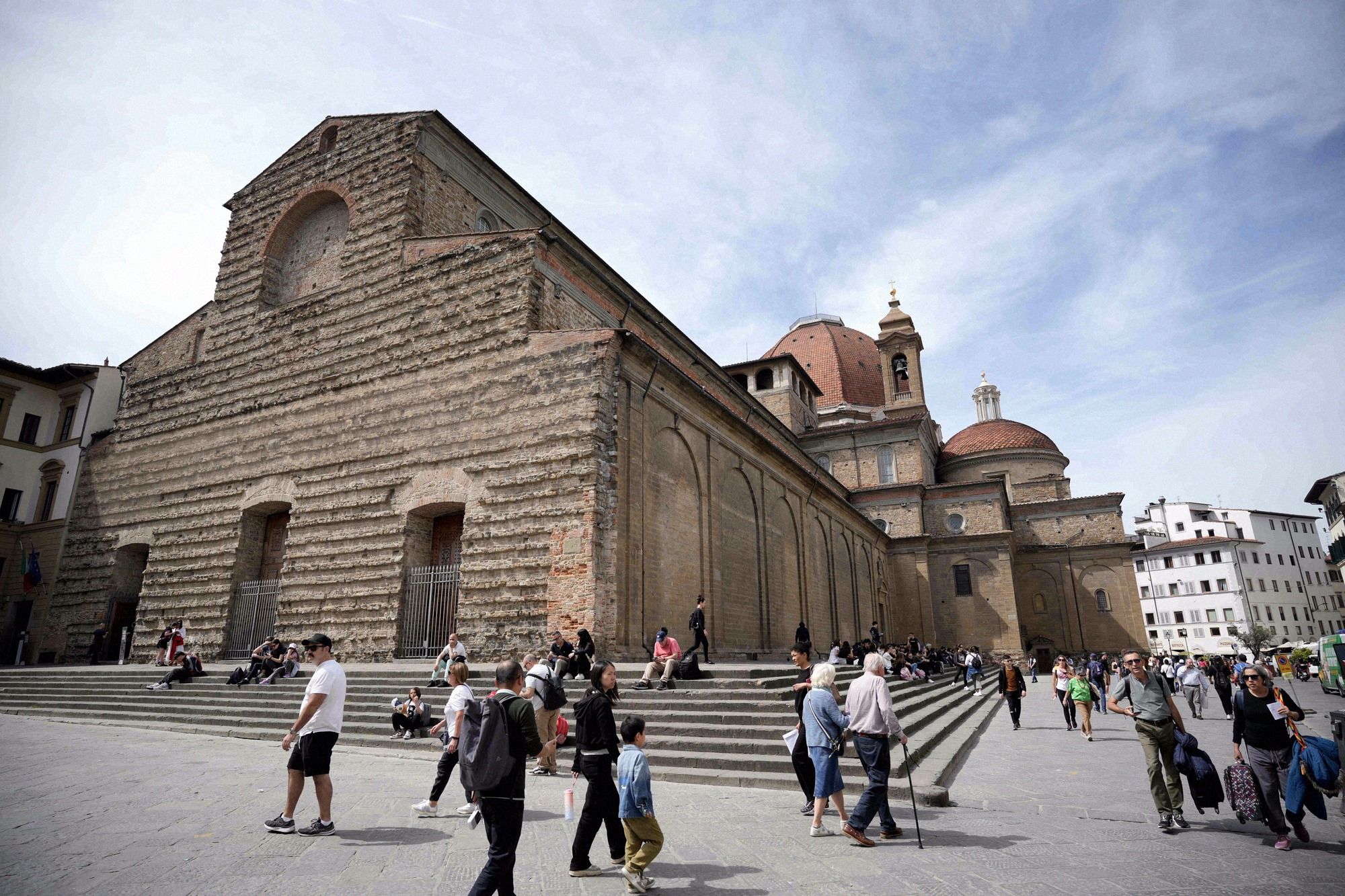 Die unverkleidete Sandstein-Fassade der Basilika San Lorenzo mit der Medici-Kuppel rechts und belebtem Vorplatz voller Touristen.
